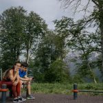 A couple in sports clothing sitting on a bench outdoors, surrounded by trees and mountains.